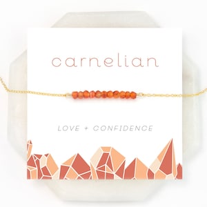 May include: A gold chain necklace with a carnelian stone bar pendant. The card reads "carnelian" and "LOVE + CONFIDENCE".