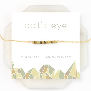 May include: A gold chain necklace with a row of green cat's eye stones. The card reads "cat's eye" and "stability + generosity".