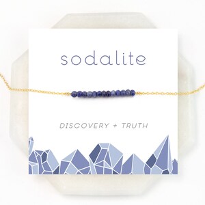 Op de afbeelding: Een gouden ketting met een staaf van blauwe sodaliet kralen. De kaart heeft de tekst "sodalite" en "ontdekking + waarheid".