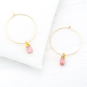 Pode incluir: Um par de brincos de argola dourados com pingentes de pedra rosa. Os brincos são simples e elegantes, perfeitos para o uso diário.