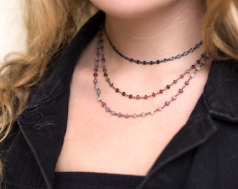 May include: A layered necklace with three strands of small, colorful beads. The top strand is a delicate chain with a black finish. The necklace is worn by a person with long, wavy hair.