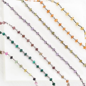 May include: A close-up of several strands of black chain with colourful gemstone beads. The beads are in shades of green, purple, pink, and orange.