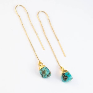 Puede incluir: Un par de pendientes colgantes dorados con colgantes de piedra turquesa. Los pendientes tienen un diseño de cadena delicada y una parte superior curvada que atraviesa la oreja. Las piedras turquesas tienen una forma natural e irregular.