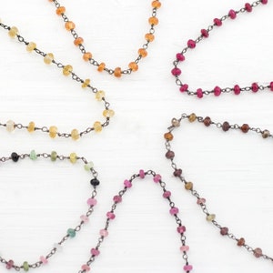 Op de afbeelding: Vijf kettingen met verschillende gekleurde edelstenen en zilveren schakels. De kettingen liggen op een wit oppervlak.