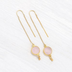 Puede incluir: Un par de pendientes de oro con detalles de piedras preciosas rosas. Los pendientes tienen una cadena delicada con una pequeña perla dorada en el extremo.