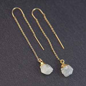 May include: A pair of gold-colored threader earrings. Each earring features a delicate chain that extends into a curved hook. At the end of each chain hangs a small, irregular-shaped, light-colored stone with gold-colored accents.