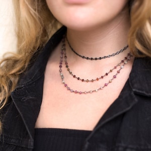 Może przedstawiać: Wielowarstwowy naszyjnik z trzema rzędami małych, kolorowych koralików. Górny rząd to delikatny łańcuszek z czarnym wykończeniem. Naszyjnik nosi osoba z długimi, falującymi włosami.