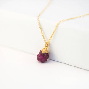 Puede incluir: Un collar de cadena de oro con un colgante de rubí en bruto. El rubí es de color rojo intenso y tiene una textura rugosa y natural. El colgante está unido a la cadena con un aro de oro.
