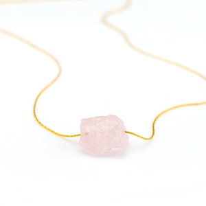 May include: A delicate gold chain necklace with a single, large, light pink, raw rose quartz crystal pendant.