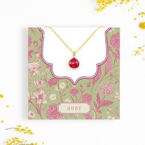 May include: A gold necklace with a red ruby gemstone pendant on a floral patterned card with the word "RUBY" printed on it.