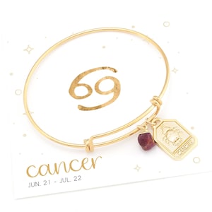 May include: Gold bangle bracelet with a charm featuring the Cancer zodiac symbol and a red gemstone. The bracelet is on a white card with the text "cancer" and "JUN. 21 - JUL. 22".