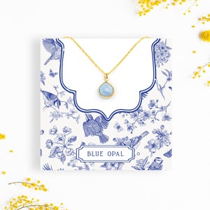 May include: A delicate gold necklace with a light blue teardrop-shaped opal pendant. The necklace is displayed on a white card with a blue and white floral pattern and the text "BLUE OPAL".
