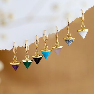 May include: A set of seven gold hoop earrings with a variety of gemstone pyramid charms. The charms are in shades of purple, green, black, blue, white, and pink.