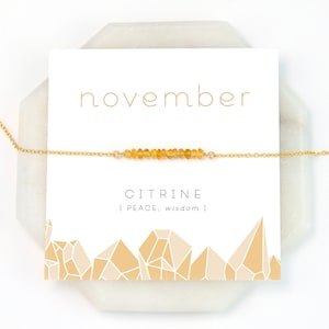 May include: A gold chain necklace with a bar of citrine stones. The card reads "november" and "CITRINE | PEACE; wisdom |"