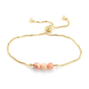 Könnte beinhalten: Ein goldfarbenes, verstellbares Armband mit drei rosa Edelsteinperlen. Das Armband hat eine feine Kette und einen Schiebeverschluss für eine individuelle Passform. Die Edelsteine haben eine zarte rosa Farbe.