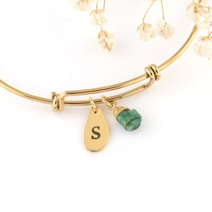 Könnte beinhalten: Goldenes Armreif-Armband mit einem kleinen Smaragd-Stein-Anhänger und einem goldenen Anhänger mit dem eingravierten Buchstaben 'S'.