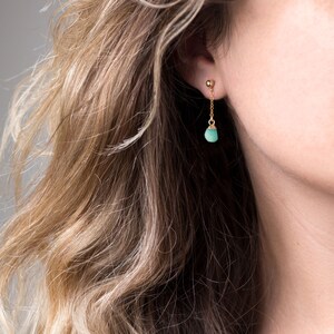 May include: A pair of gold earrings with a small, teardrop-shaped turquoise gemstone dangling from each earring.