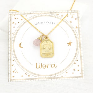 May include: Gold necklace featuring a Libra zodiac charm and a pink crystal pendant. The necklace is presented on a white card with gold stars and the text "SEPT. 23 - OCT 22" and "Libra".