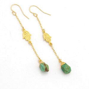 Puede incluir: Pendientes de aro dorados con un dije de mano de hamsa y una gota de piedra turquesa.