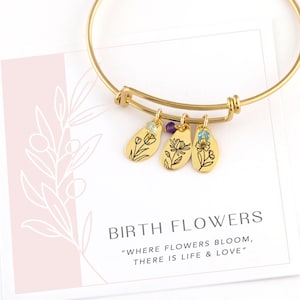Puede incluir: Pulsera de brazalete de oro con tres dijes con diseños florales grabados y pequeñas piedras preciosas. La tarjeta dice "Birth Flowers" y "Where flowers bloom, there is life & love."