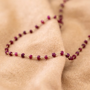 May include: A delicate gold chain necklace with small, deep red ruby beads. The necklace is draped on a soft, brown fabric.