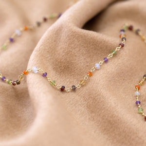 Może przedstawiać: Złoty łańcuszek z małymi, kolorowymi koralikami z kamieni szlachetnych. Koraliki są w odcieniach pomarańczowego, czerwonego, fioletowego, zielonego i niebieskiego.