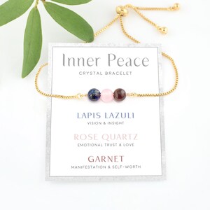Peut inclure: Bracelet ajustable en or avec trois perles de pierres précieuses : lapis-lazuli, quartz rose et grenat. La carte indique "Inner Peace Crystal Bracelet" et liste les pierres avec leurs significations associées.