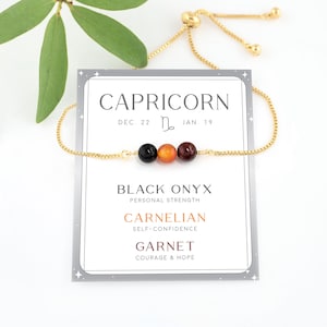 May include: A gold chain bracelet with three gemstone beads: black onyx, carnelian, and garnet. The card reads "Capricorn Dec. 22 - Jan. 19" and lists the stone meanings: Black Onyx - Personal Strength, Carnelian - Self-Confidence, Garnet - Courage & Hope.