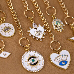 May include: A set of gold chain necklaces with charms featuring white enamel and blue and green accents. The charms are shaped like hearts, circles, and hands with an evil eye design.