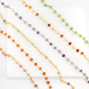 Może przedstawiać: Zestaw sześciu złotych łańcuszków z koralikami z kamieni szlachetnych w różnych kolorach. Łańcuszki ułożone są w rzędzie na białym tle. Kamienie szlachetne są w odcieniach pomarańczowego, fioletowego, zielonego i różowego.
