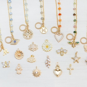 Op de afbeelding: Een verzameling gouden bedelarmbanden met verschillende bedels, waaronder een kompas, een madeliefje, een bij, een vlinder, een zeester, een kruis, een hart, een hand, een lotusbloem, een oog, een wolk, een zon en de letter M.