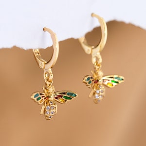 May include: Gold hoop earrings with a dangling bee charm. The bee is decorated with colorful enamel and clear rhinestones.