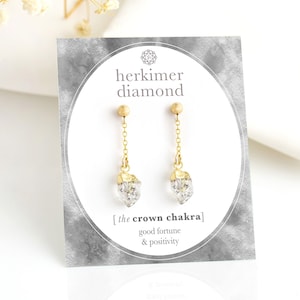 May include: Gold-colored Herkimer diamond earrings with a clear, crystal-like appearance. The earrings hang from a gold chain and are displayed on a card with the text "herkimer diamond" and "the crown chakra" with "good fortune & positivity".