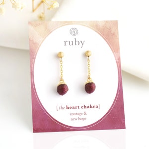 Puede incluir: Pendientes de oro con un colgante de rubí rojo. Los pendientes están en una tarjeta blanca con el texto "ruby" y "[the heart chakra] courage & new hope".
