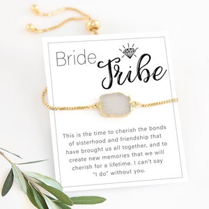 Op de afbeelding: Een gouden kettingarmband met een witte stenen bedel en een kaart met de tekst "Bride Tribe". De kaart bevat een boodschap over het koesteren van de banden van zusterschap en vriendschap.