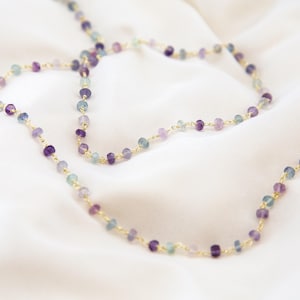 Puede incluir: Un collar largo de cadena dorada con pequeñas cuentas facetadas en tonos de morado, azul y verde.