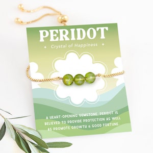 Puede incluir: Un brazalete ajustable de oro con tres piedras de peridoto verdes. El brazalete viene con una tarjeta que dice "Peridoto, Cristal de la Felicidad" y describe el significado de la piedra.