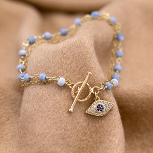 May include: A gold chain bracelet with blue and white beads and a gold evil eye charm. The bracelet is on a brown fabric background.