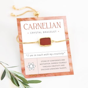May include: A gold adjustable bracelet with a carnelian stone. The card reads "CARNELIAN + CRYSTAL BRACELET + "I am in touch with my creativity" STONE OF CONFIDENCE AND MOTIVATION. EXPRESS YOURSELF THROUGH CREATIVITY AND DISCOVER UNKNOWN TALENTS."