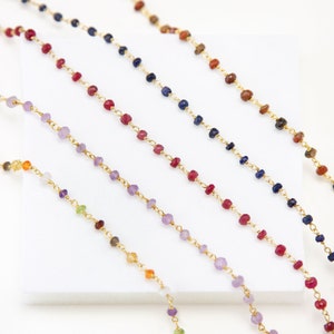 Puede incluir: Cinco cadenas de oro con cuentas de piedras preciosas de diferentes colores. Las cadenas están dispuestas en fila sobre una superficie blanca. Las piedras preciosas son amatista, rubí, zafiro, ojo de tigre y una mezcla de varios colores.