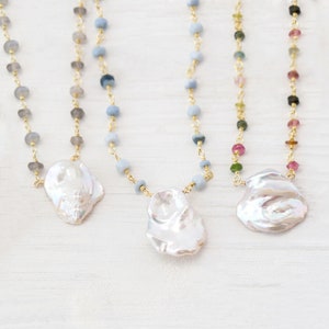 May include: Four necklaces with different colored beads and a large, white baroque pearl pendant. The necklaces are on a white wooden surface.