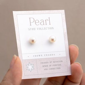 Op de afbeelding: Een paar witte parel oorbel studs op een witte kaart met de tekst "Pearl Stud Collection" en "Crown Chakra". De kaart bevat ook de tekst "Chakra of Intuition, Sense of Purpose, and Connection".