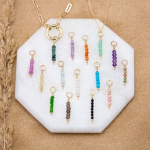 May include: A collection of 16 gold charm pendants with different colored gemstone beads. The charms are arranged on a white marble surface.