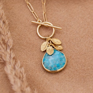 Op de afbeelding: Een gouden ketting met een turquoise stenen hanger en kleine gouden blaadjescharms. De ketting heeft een toggle sluiting.