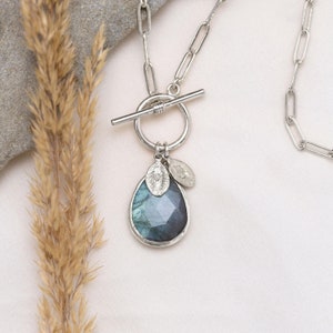 Puede incluir: Un collar de plata con cierre de palanca y un colgante en forma de lágrima con una piedra de labradorita azul. El colgante está adornado con dos pequeños amuletos de plata.