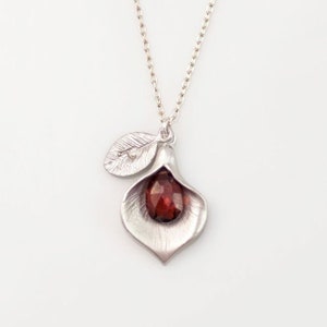Peut inclure: Un collier en argent avec un pendentif en forme de fleur avec une pierre précieuse rouge au centre. Le pendentif a un petit charme de feuille avec un petit diamant.