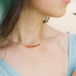 May include: A gold chain necklace with a row of small, orange carnelian beads. The necklace is 15 inches long and is shown on a model wearing a light blue top.
