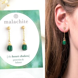 May include: Malachite earrings with a gold chain and green gemstone. The earrings are on a card with the text "malachite" and "the heart chakra" with the words "protection & balance" below.