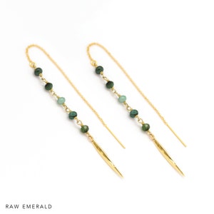 K&ouml;nnte beinhalten: Goldene Ohrh&auml;nger mit gr&uuml;nen Smaragdperlen und einem goldenen Stachelanh&auml;nger. Die Ohrringe werden vor wei&szlig;em Hintergrund gezeigt. Der Text "RAW EMERALD" befindet sich am unteren Bildrand.