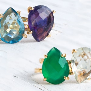 May include: Two gold rings with teardrop-shaped gemstones. The top ring features a light blue gemstone and a purple gemstone. The bottom ring features a green gemstone and a light green gemstone.